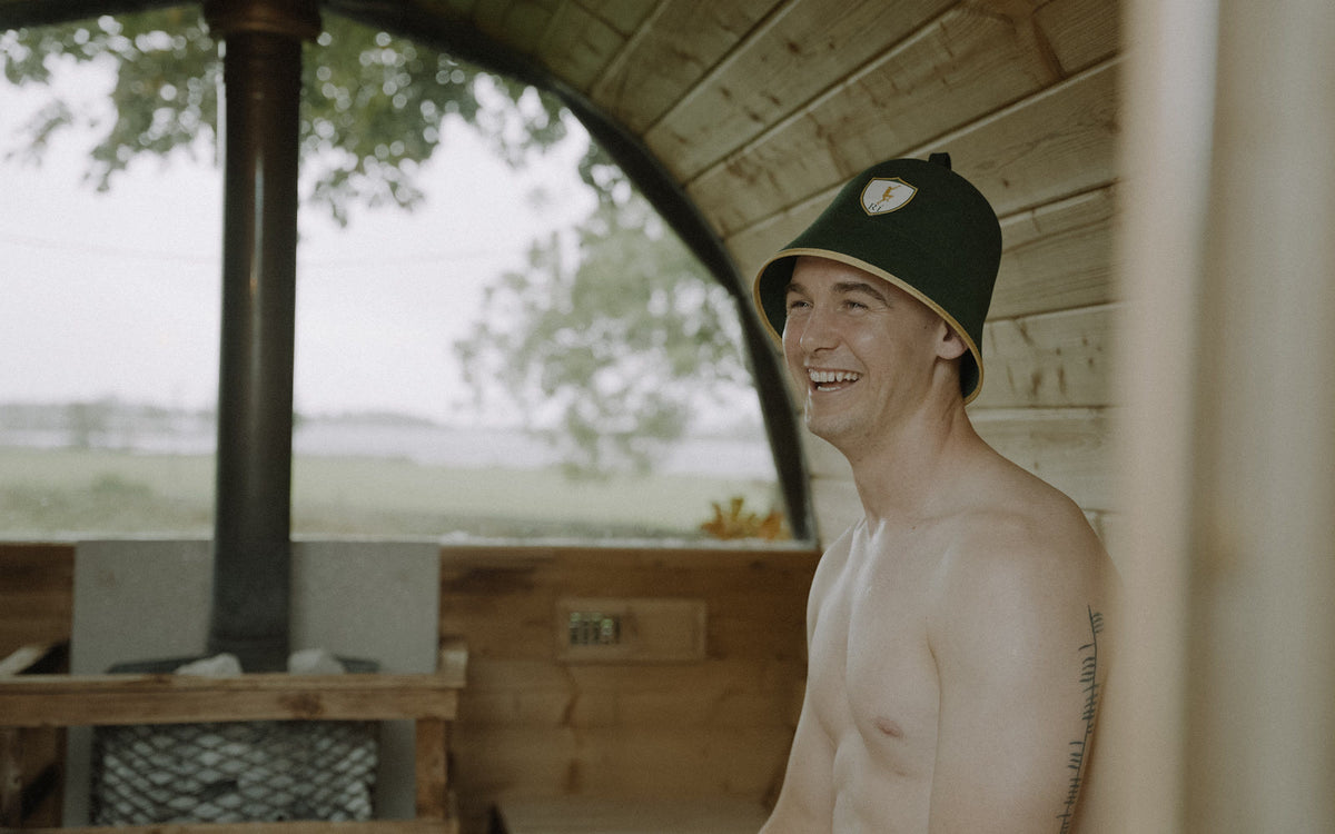 A man wearing the Rí Sauna Crown inside a barrel sauna, lake behind