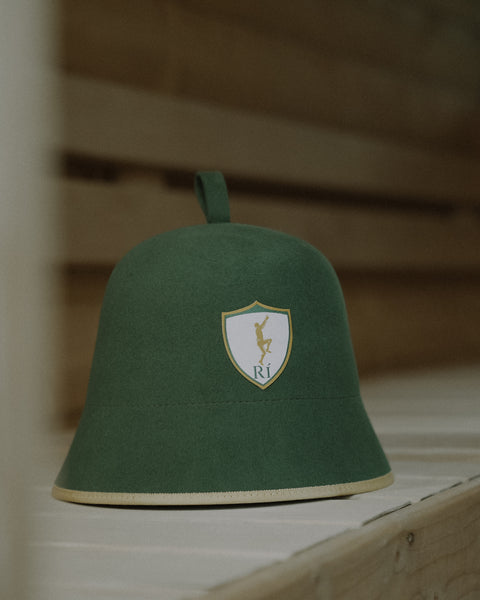 Rí Sauna Crown, deep green merino wool sauna hat on a sauna bench, shield crest facing forward